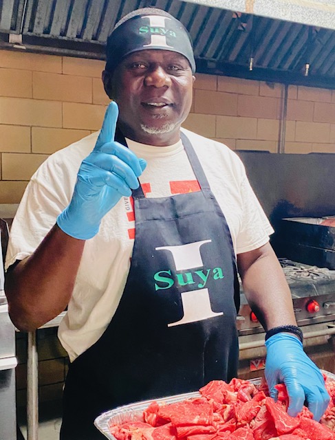 A person in a ‘Suya’ apron holds a tray of red meat, ready for grilling